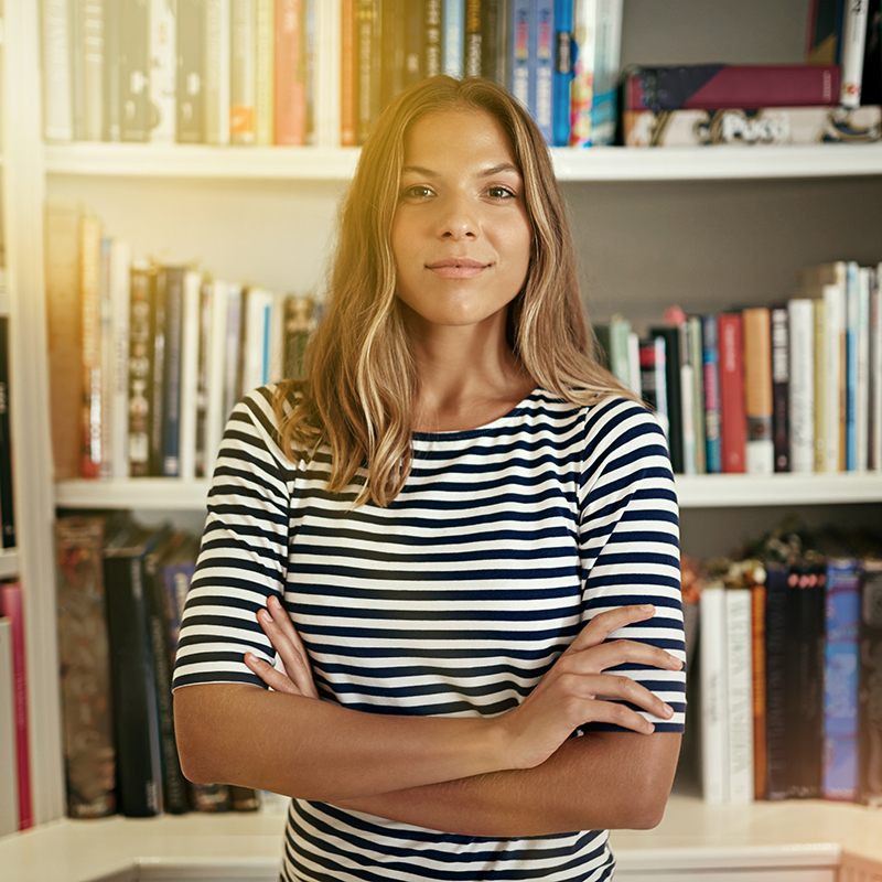 Woman, arms crossed and portrait by bookshelf with smile, pride